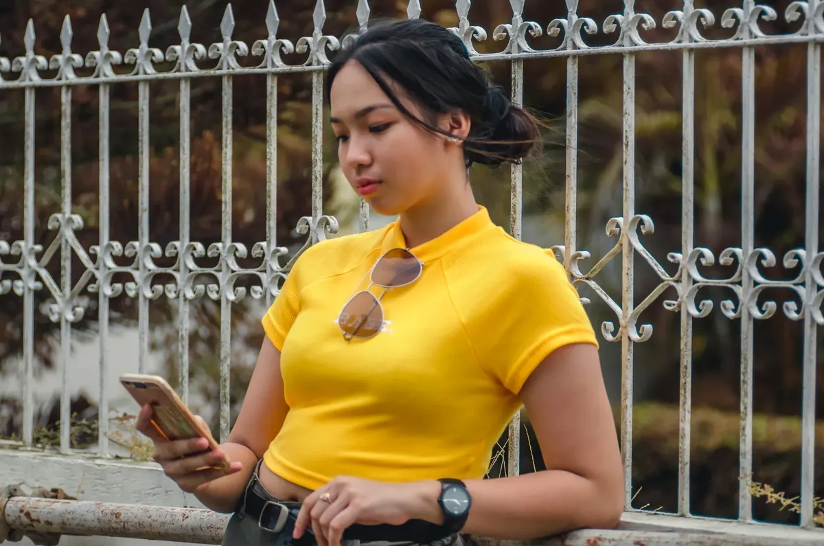 Young woman in bright yellow top using smartphone while leaning against a decorative fence.