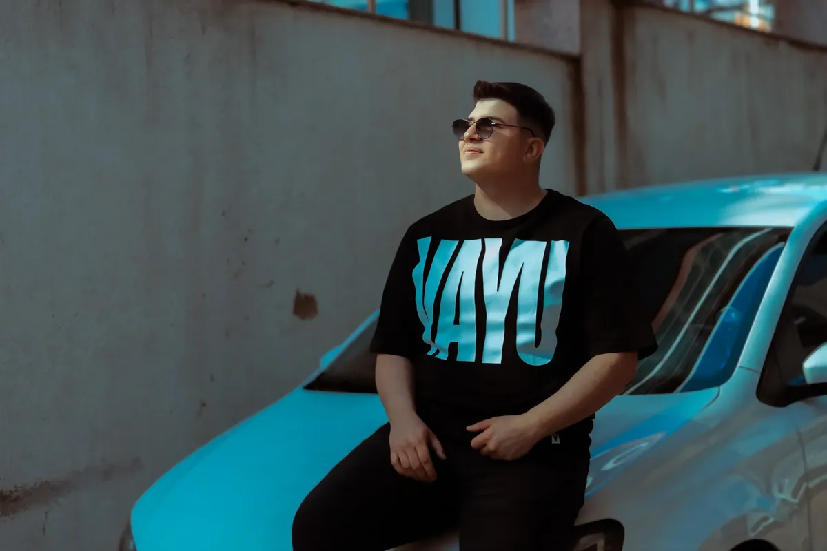 A man wearing sunglasses and a t-shirt sits on a car hood in a sunny urban environment.