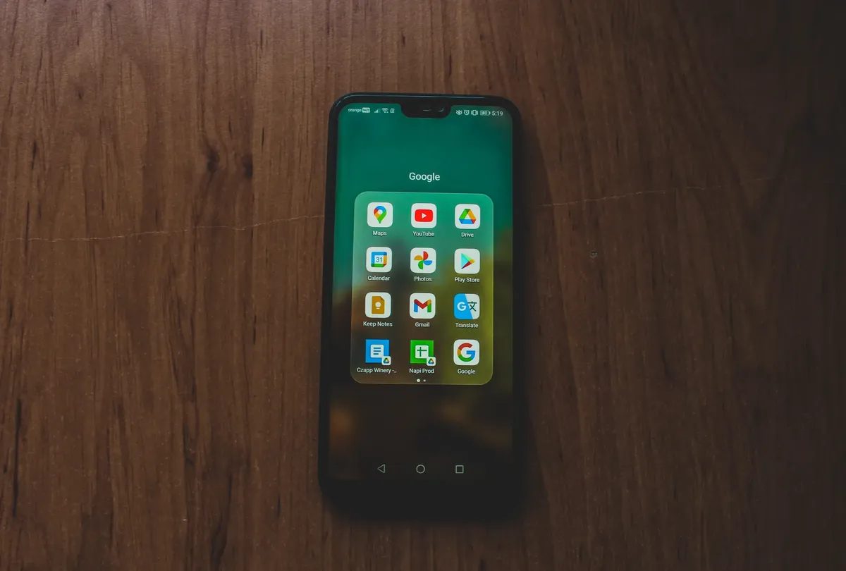 A smartphone showcasing various Google apps on a wooden table.